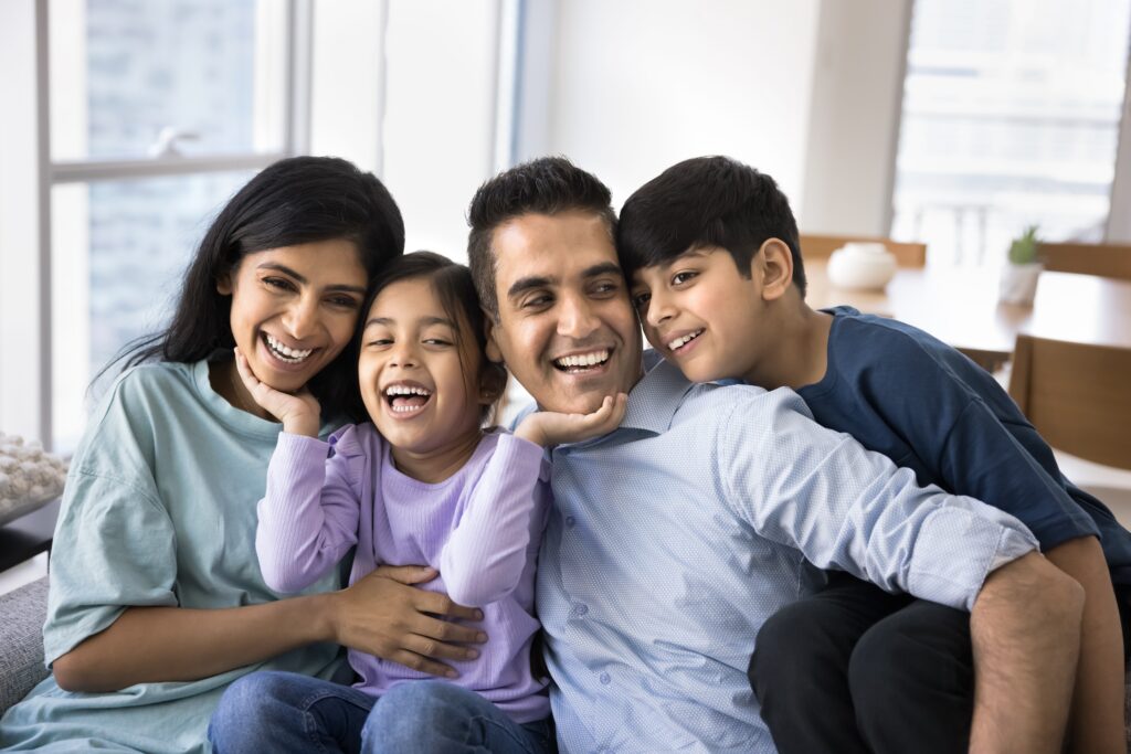 Happy excited parents and two kids having fun at home, sitting close on couch, hugging, laughing, holding younger child in arms, enjoying homey family leisure together